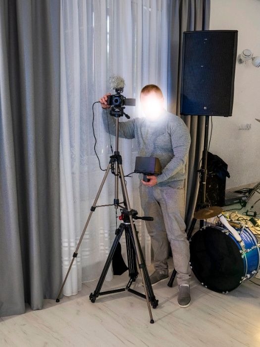Osoba ustawia kamerę z mikrofonem na statywie, obok głośnik i perkusja, przygotowania do nagrania.