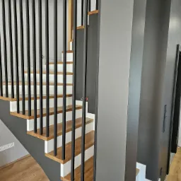 Nowoczesne schody z drewnianymi stopniami, białymi podstopnicami i czarną, metalową balustradą na tle szarej ściany. Widoczne detale wykończenia wnętrza.