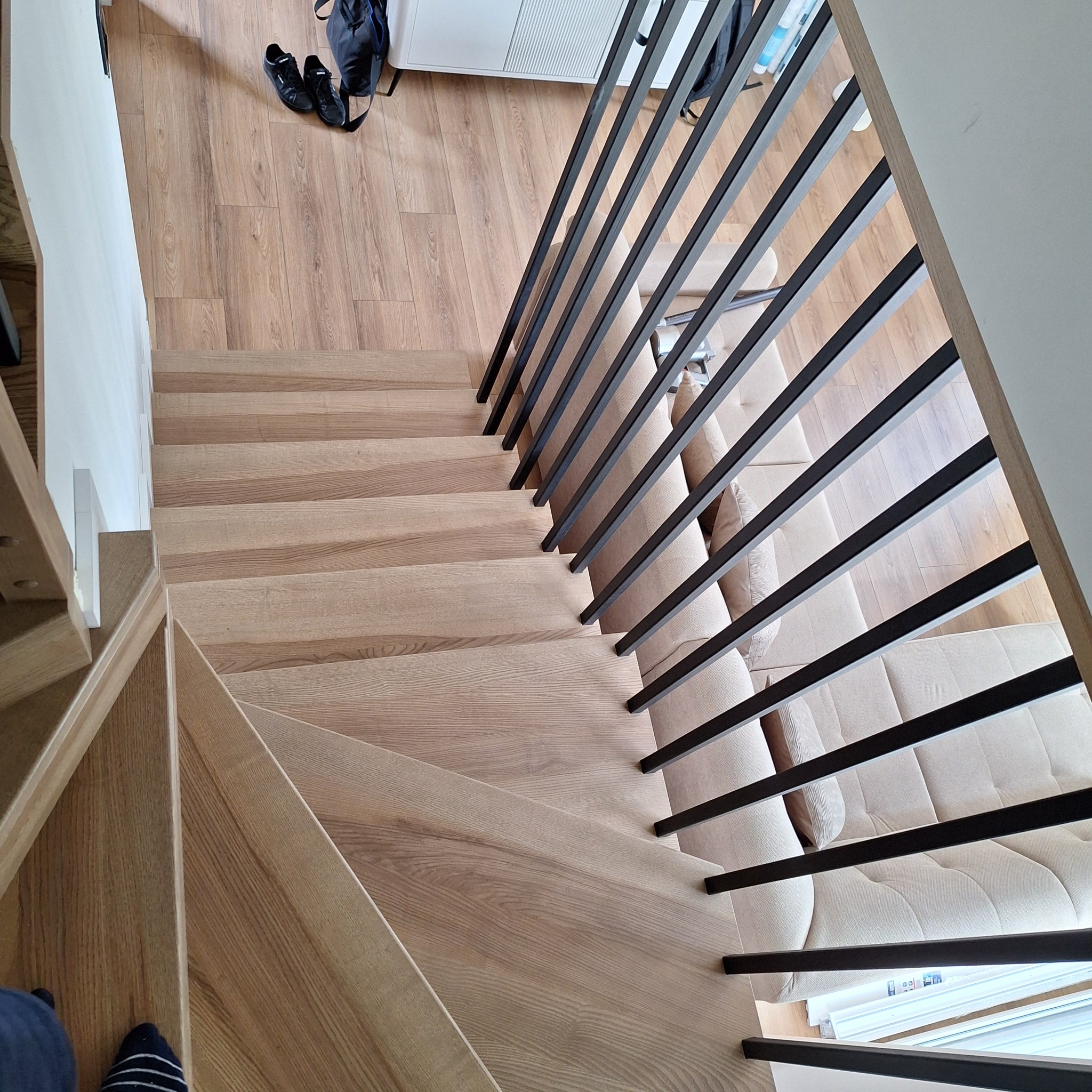 Jasne schody drewniane z czarną, nowoczesną balustradą widziane z góry. Na podłodze widoczne buty i torba, co dodaje realizmu i sugeruje użytkowanie.