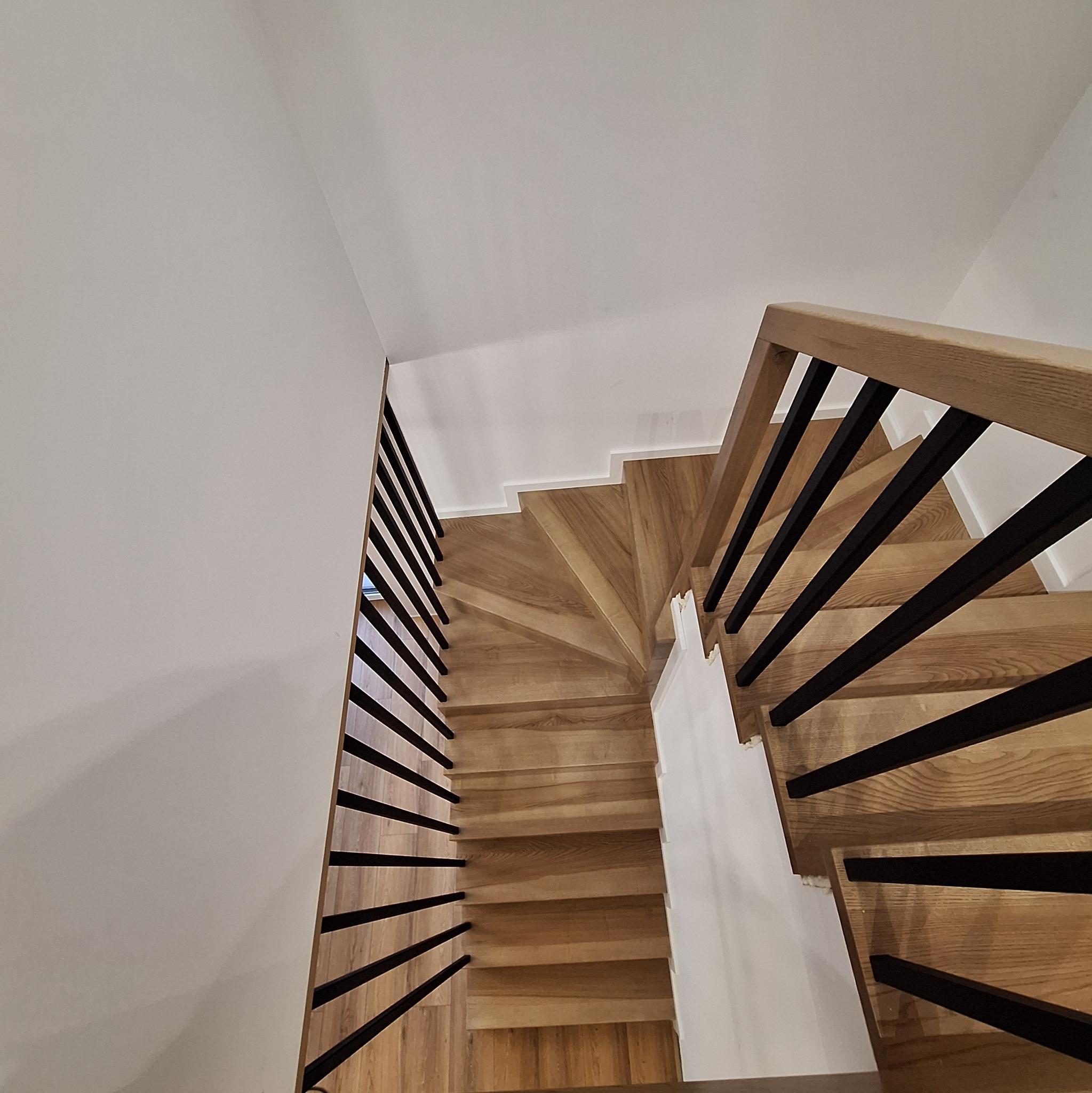 Kątowe ujęcie na drewniane schody spiralne z czarnymi, metalowymi balustradami, prowadzące w górę. Jasne drewno kontrastuje z bielą ścian i sufitu.