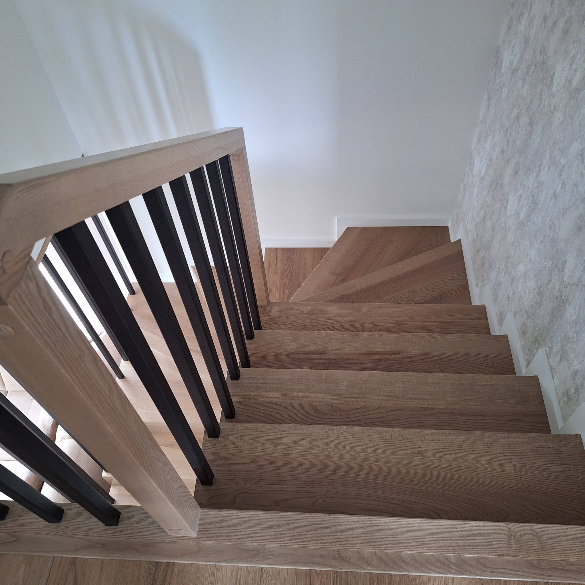 Nowoczesne schody z drewna dębowego, z czarnymi, metalowymi balustradami. Widok z góry na stopnie i fragment ściany z dekoracyjnym tynkiem.