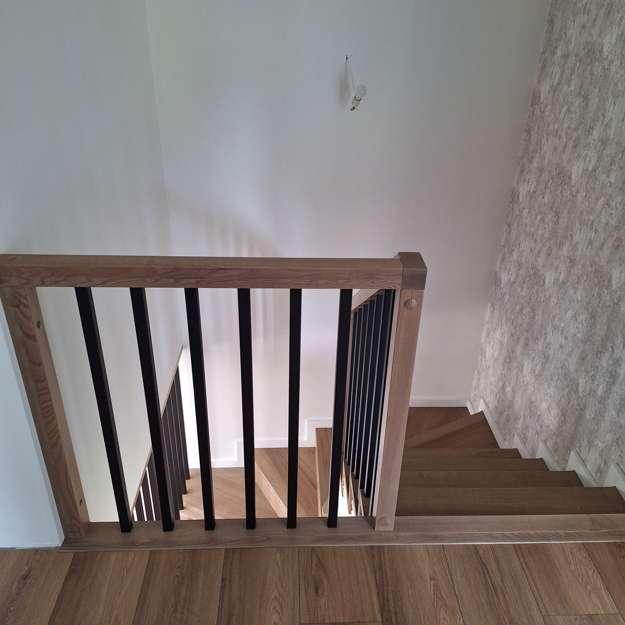Drewniane schody z czarnymi balustradami i jasną drewnianą podłogą, widok z góry. Ściana z tapetą w tle.