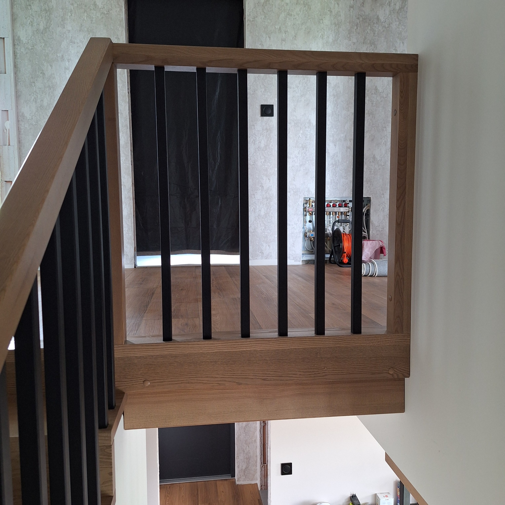 Jasne drewniane schody z czarnymi balustradami, widok z góry na fragment klatki schodowej, w tle pomieszczenie z instalacją grzewczą.