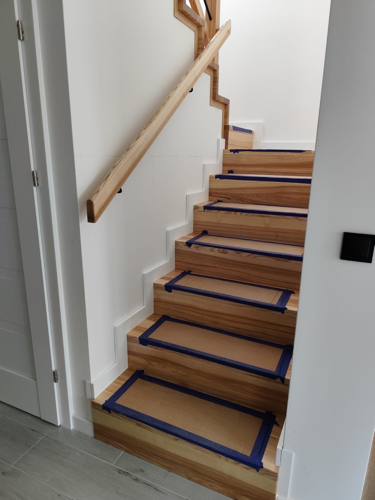 Jasne, drewniane schody z zabezpieczonymi stopniami, prowadzące w górę, z drewnianą poręczą i białymi ścianami. Widoczne zabezpieczenia na stopniach.