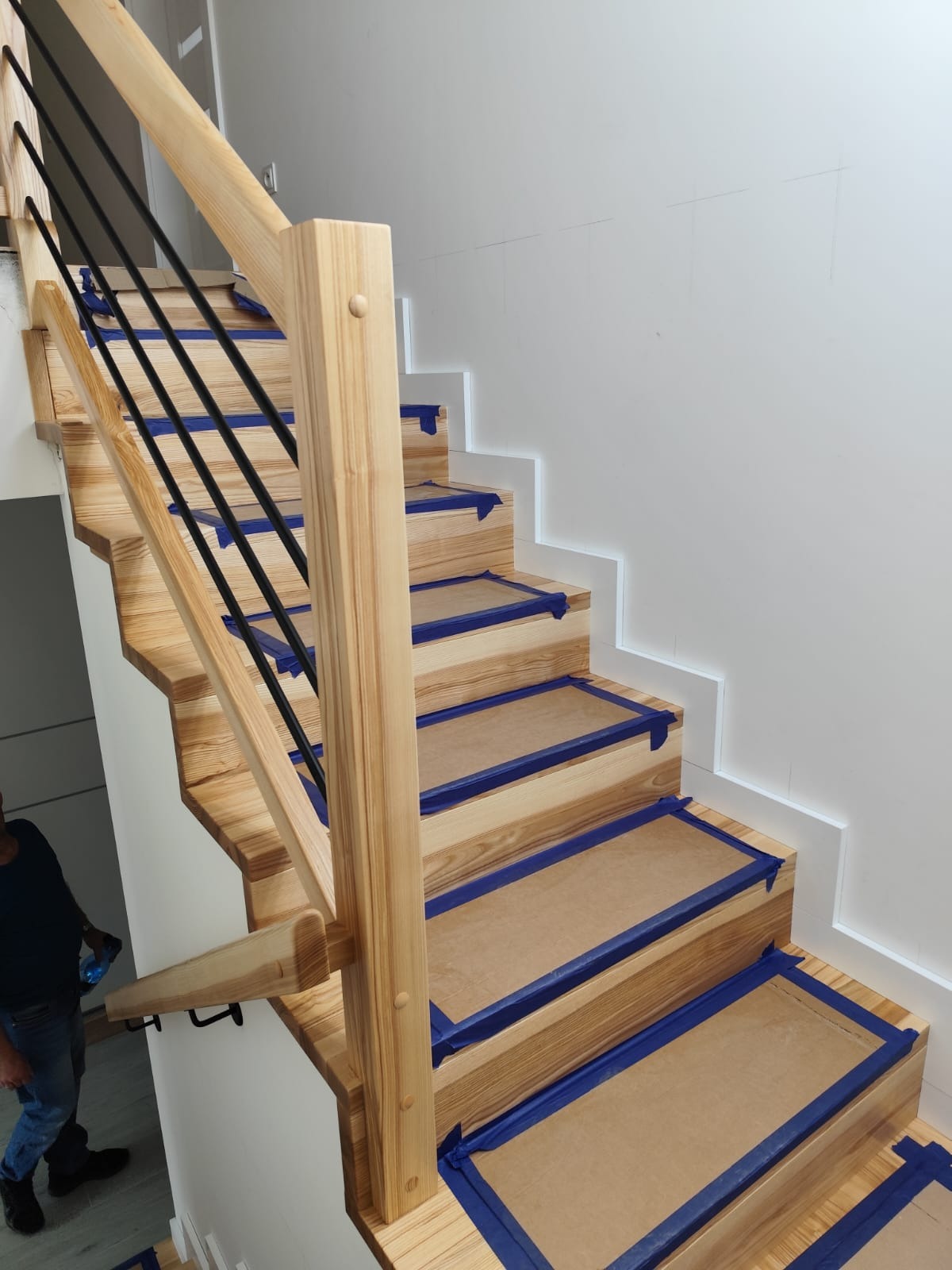 Jasne drewniane schody z czarnymi balustradami, zabezpieczone niebieską taśmą malarską i kartonem, w trakcie montażu lub wykończenia, widok z dołu.