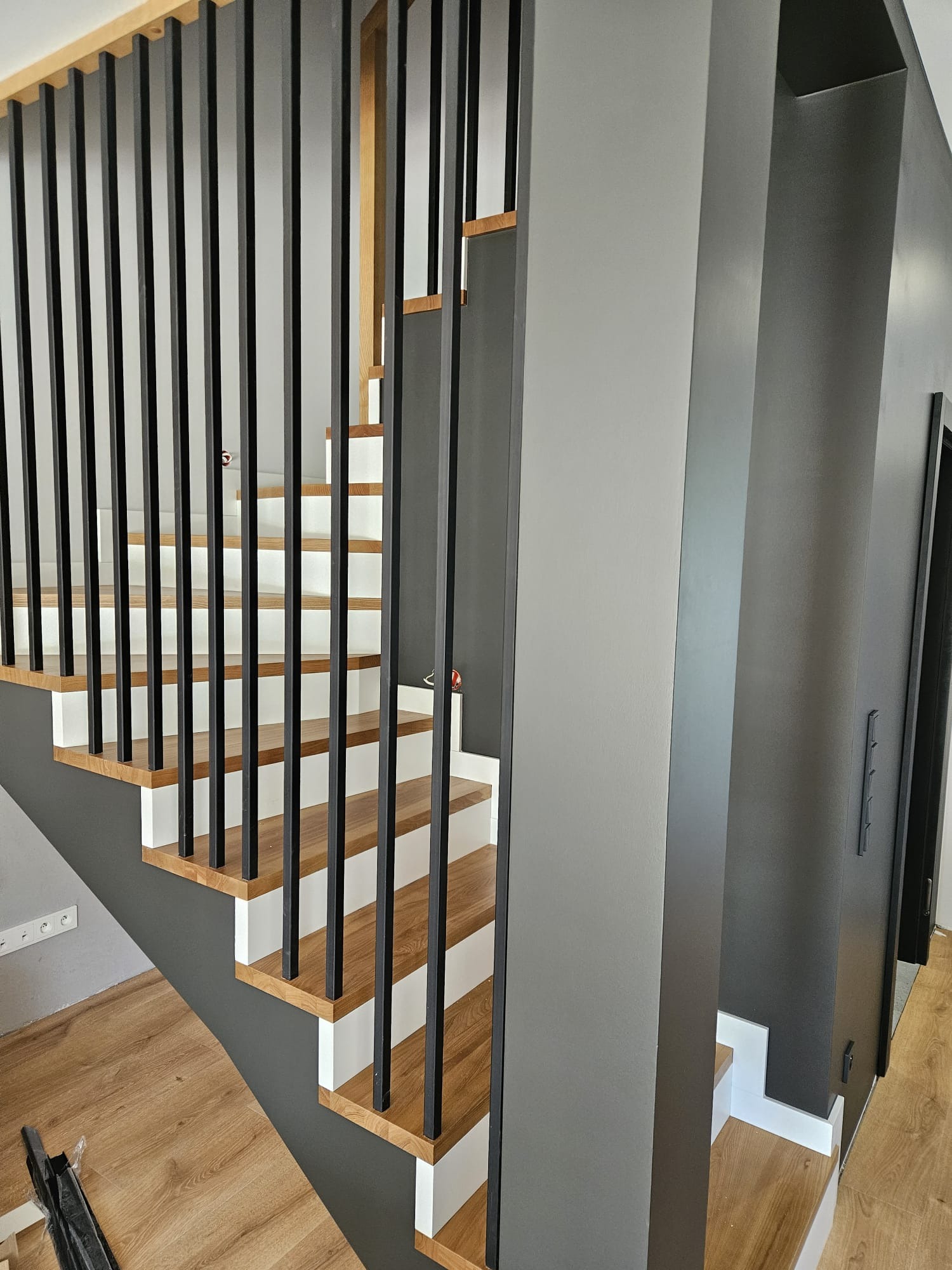 Nowoczesne schody z drewnianymi stopniami, białymi podstopnicami i czarną, metalową balustradą na tle szarej ściany. Widoczne detale wykończenia wnętrza.