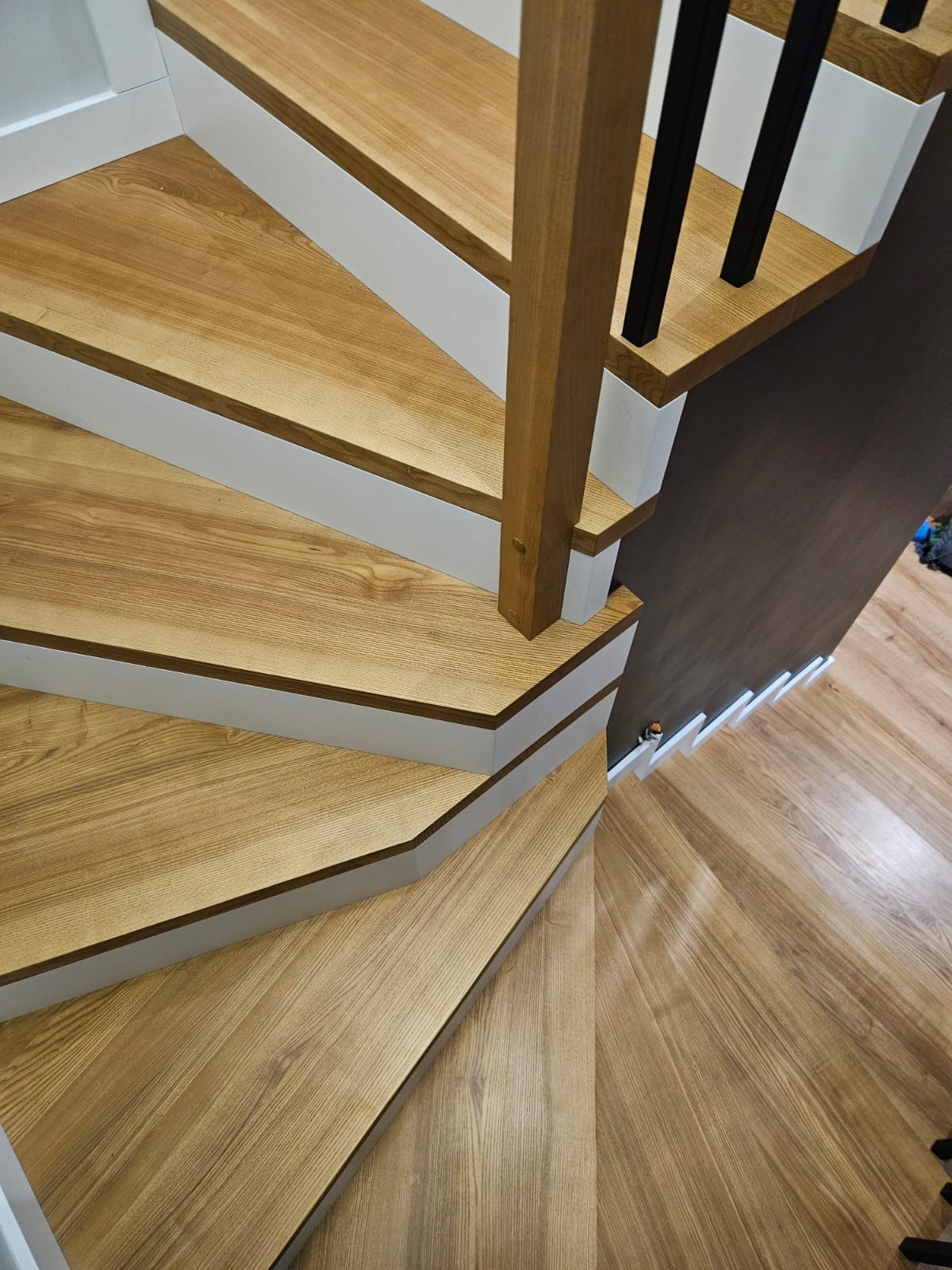 Kątowe ujęcie dębowych schodów z białymi podstopnicami i czarnymi balustradami, prowadzących w dół. Widoczne usłojenie drewna i fragment podłogi.
