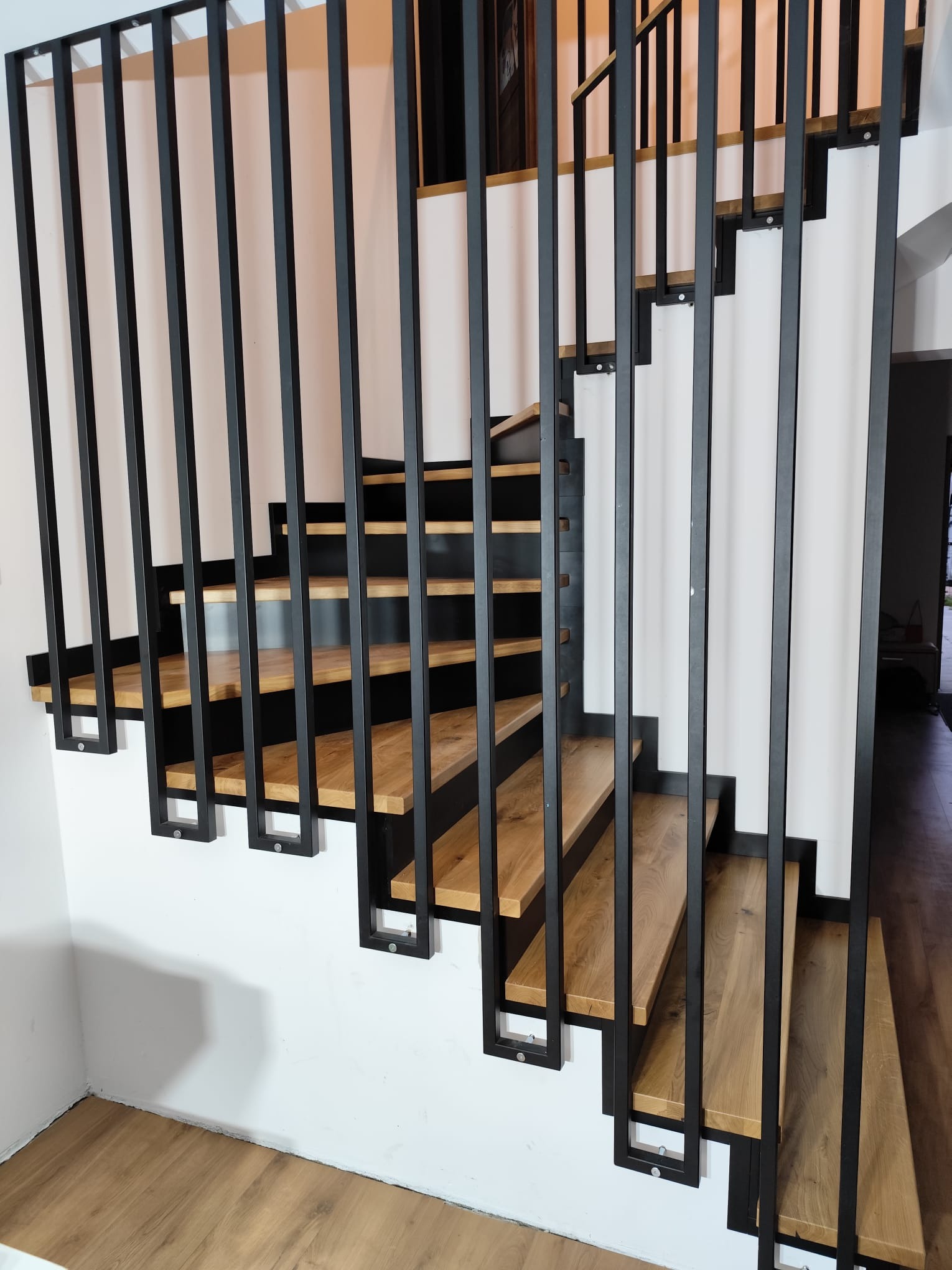 Nowoczesne schody z drewnianymi stopniami i czarną, metalową balustradą. Minimalistyczny design, jasne wnętrze, widok z dołu. Charakterystyczne mocowanie stopni.