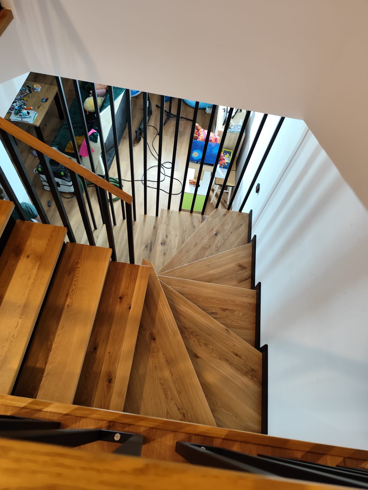 Spiralne schody z jasnego drewna z czarną balustradą, widziane z góry. Minimalistyczny design, naturalne oświetlenie, fragment pokoju dziecięcego w tle.