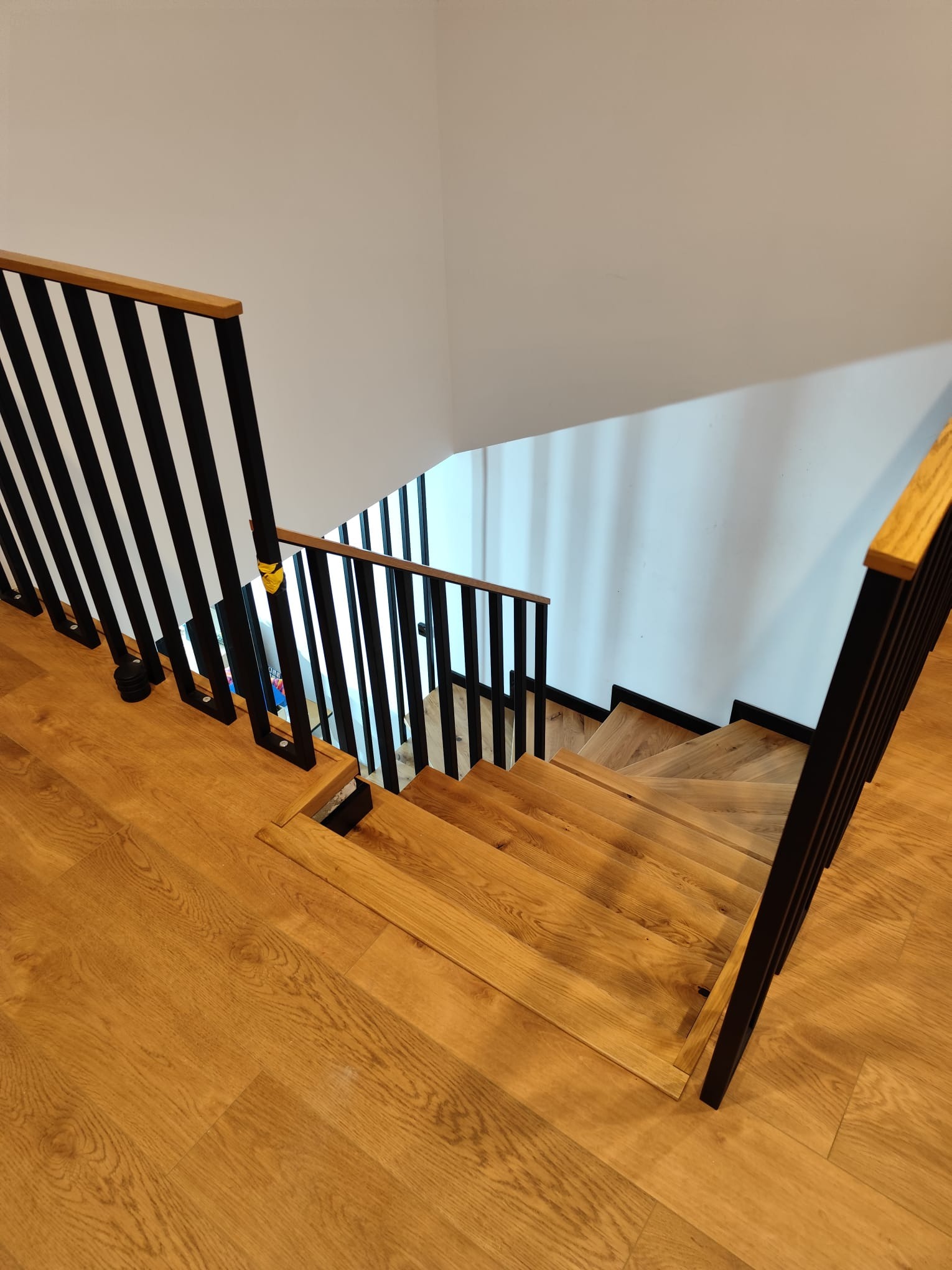 Jasne, drewniane schody z czarną, nowoczesną balustradą i drewnianym poręczą. Widok z góry na dół, podkreślający naturalne usłojenie drewna i minimalistyczny design.