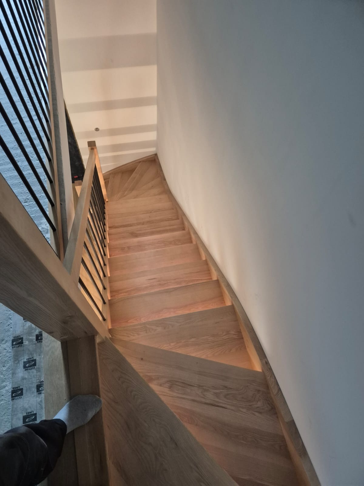 Spiralne schody z jasnego drewna z czarnymi balustradami, widok z góry, podkreślający zakrzywiony kształt i naturalne oświetlenie. Widoczny fragment stopy na dole kadru.