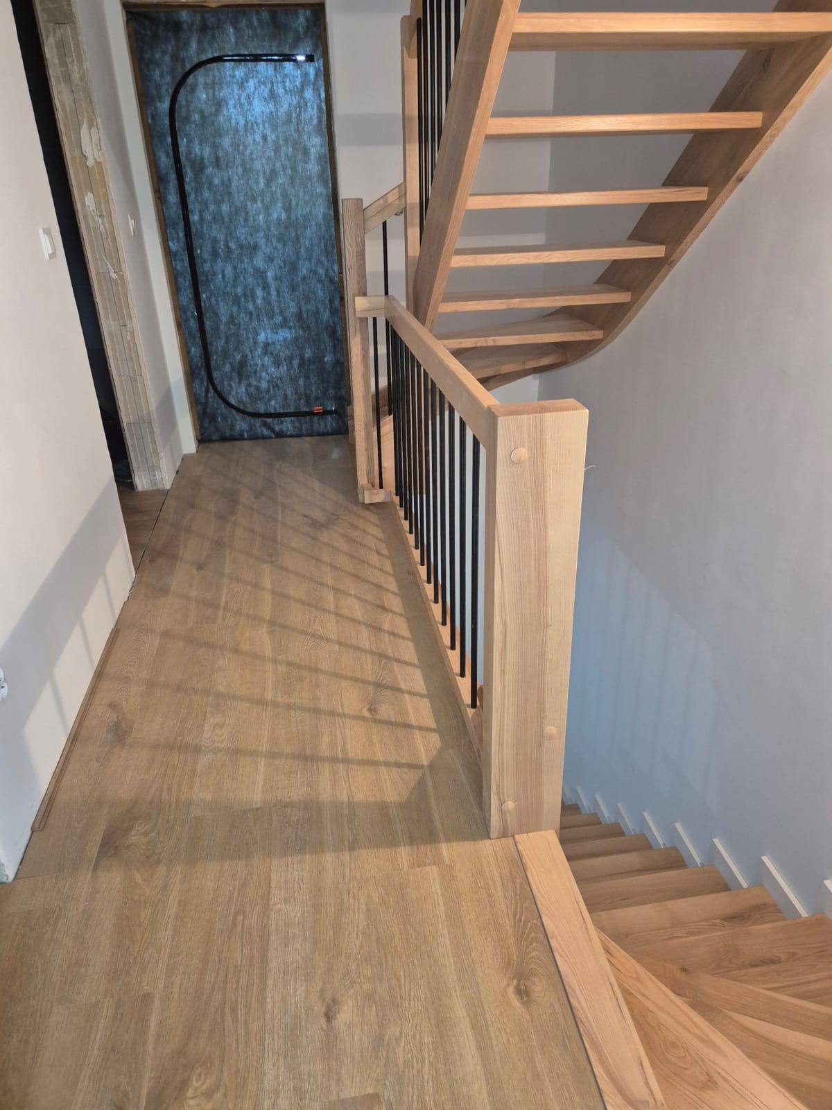 Jasne drewniane schody z czarnymi balustradami, widoczne z góry. Uwagę przyciąga nietypowe umiejscowienie drzwi na końcu korytarza.