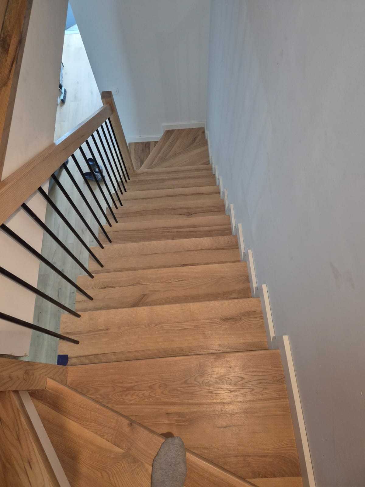 Nowoczesne, jasne stopnie z drewna z czarną balustradą, widok z góry. Fragment stopy w szarej skarpetce. Minimalistyczny design, neutralne tło ścian.