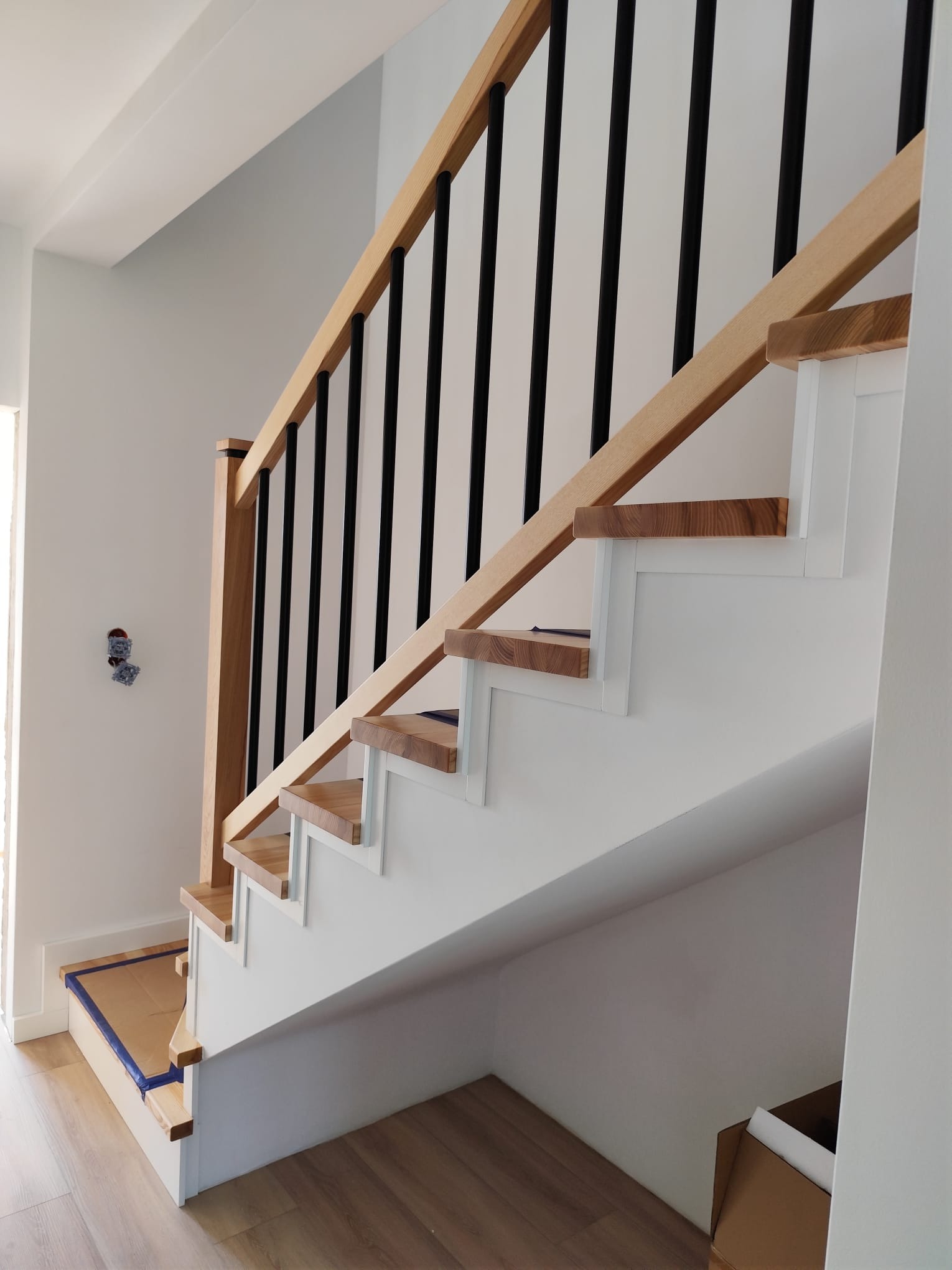 Nowoczesne schody z drewnianymi stopniami i czarną balustradą, białe boki, widok z dołu, jasne wnętrze, minimalistyczny design.