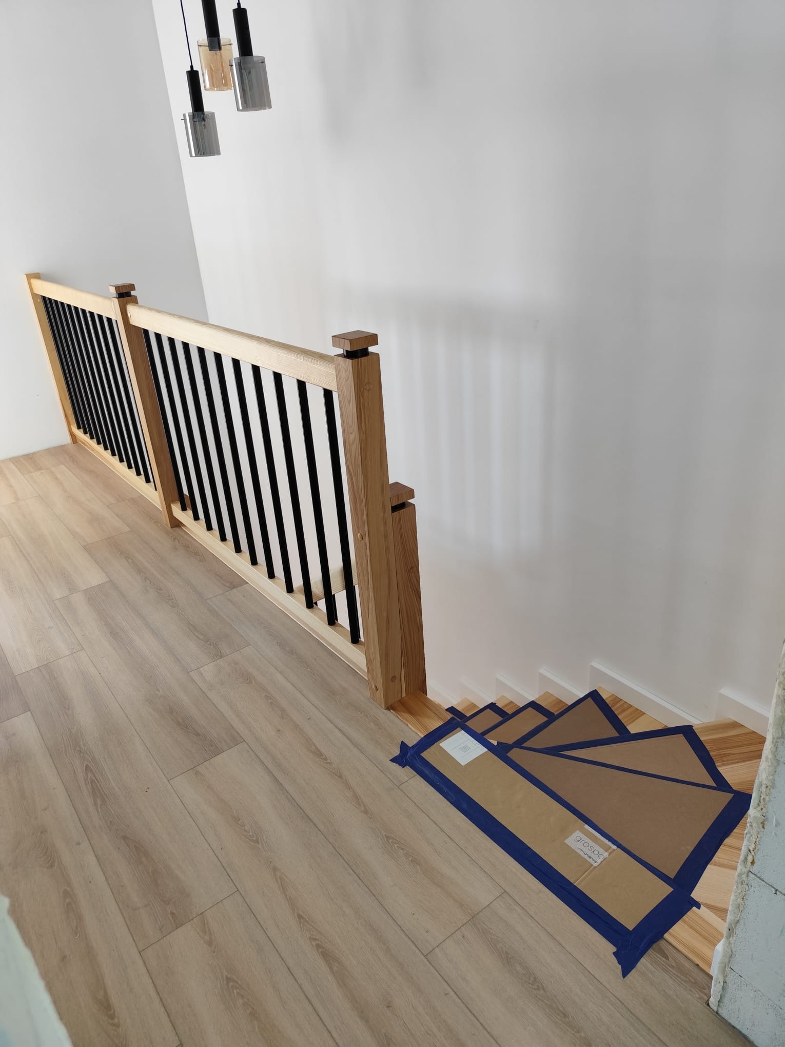 Jasne drewniane schody z czarną balustradą, zabezpieczone kartonem na czas remontu. Widok z góry, nowoczesne lampy wiszące.