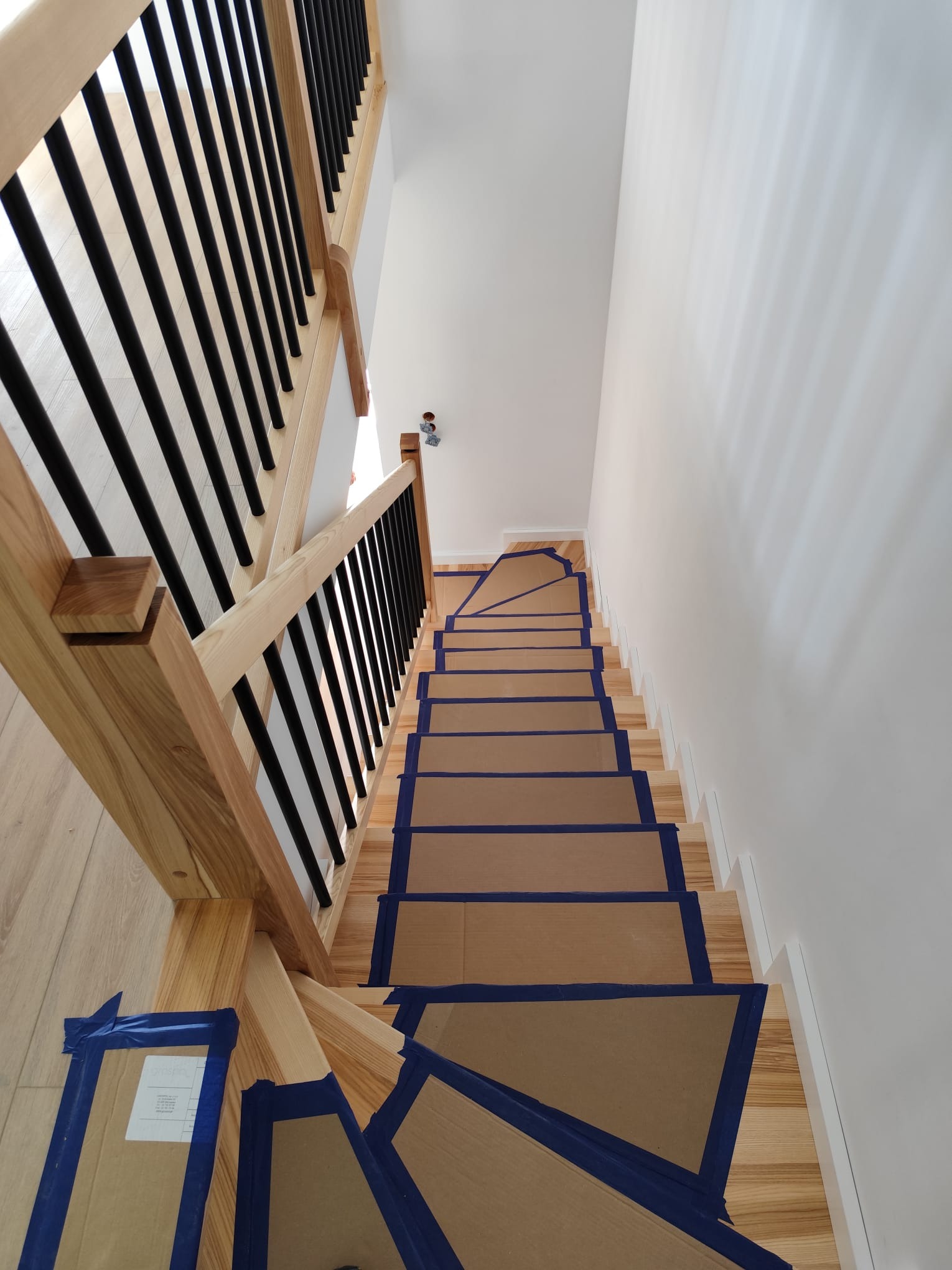 Jasne drewniane schody z czarną balustradą zabezpieczone kartonem i taśmą malarską przed malowaniem ścian. Widok z góry.
