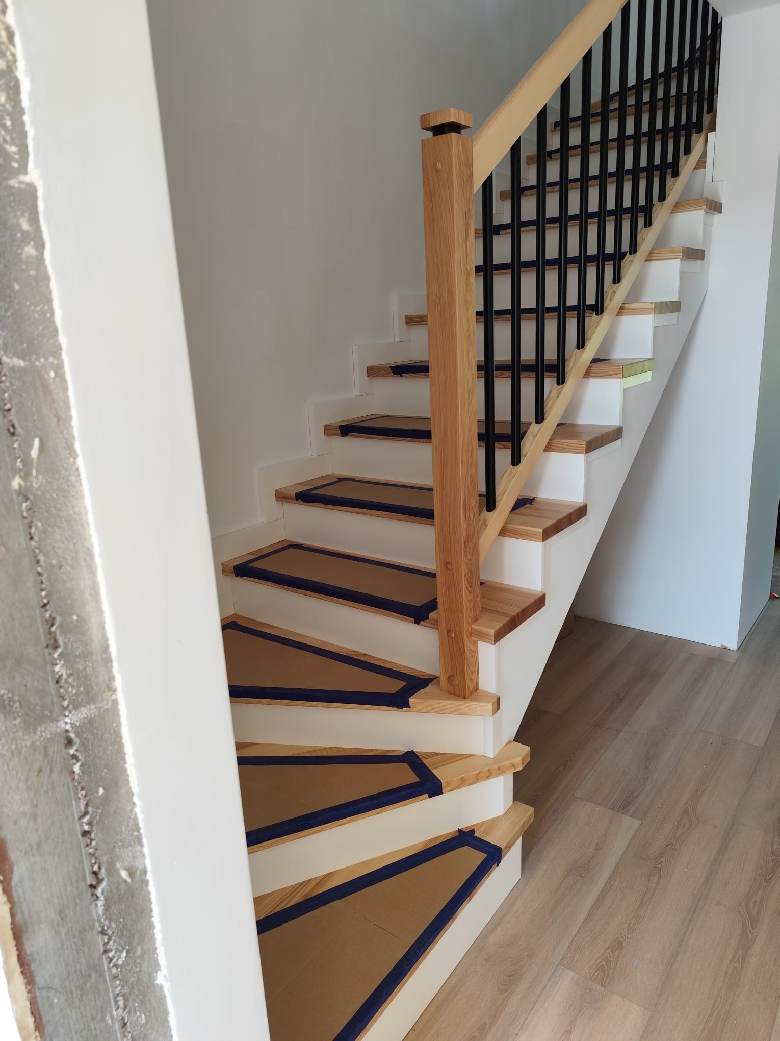 Jasne schody z drewnianymi stopniami zabezpieczonymi kartonem i niebieską taśmą, białe podstopnice, czarne metalowe barierki i drewniana poręcz. Widoczny fragment ściany w stanie surowym.