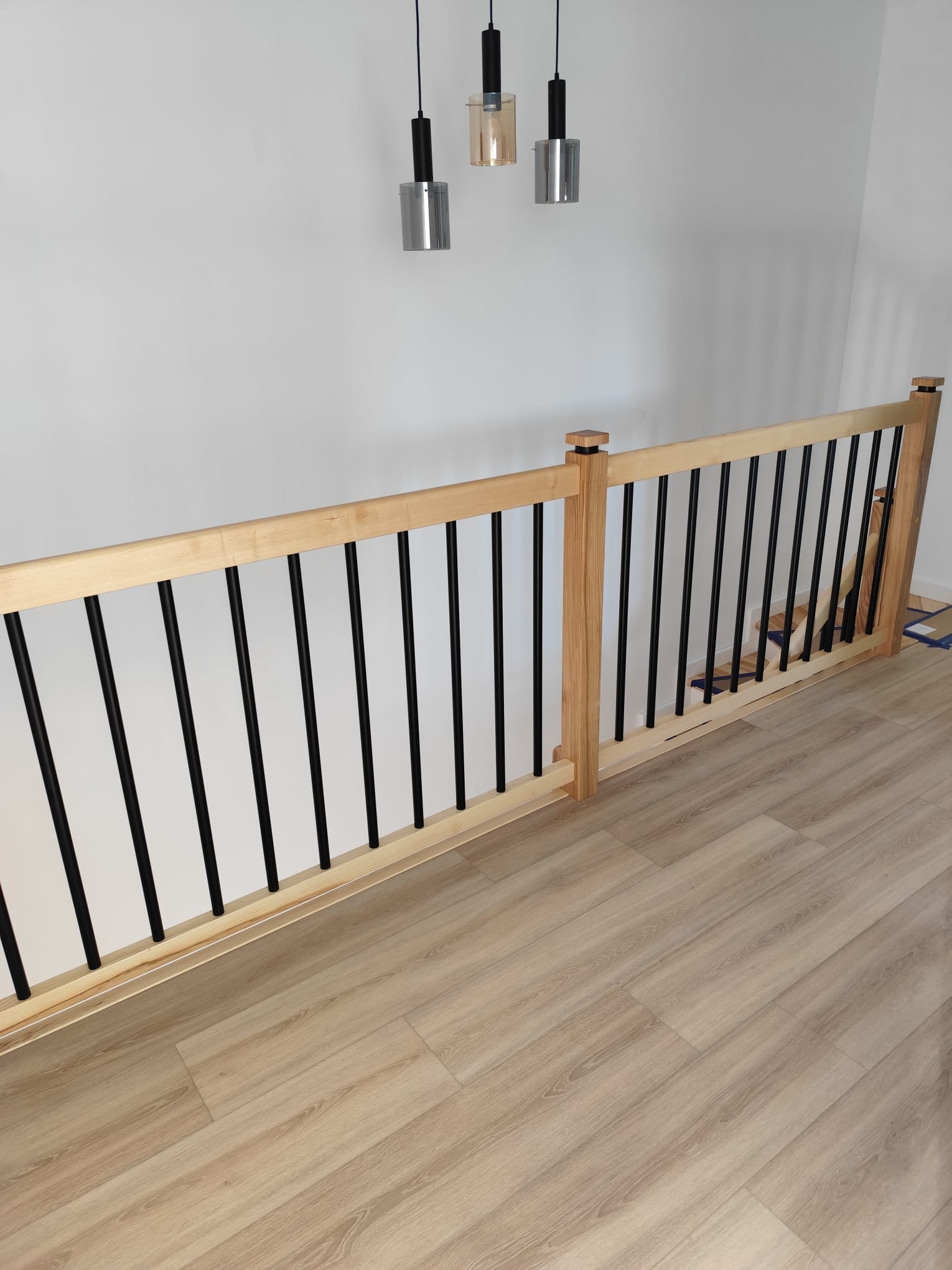 Jasne drewniane barierki schodowe z czarnymi, pionowymi szczebelkami, nowoczesne oświetlenie sufitowe, parkiet. Minimalistyczny design.