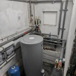 Szara pompa ciepła typu monoblok, marki Ferroli, zainstalowana w kotłowni, z widocznymi rurami w izolacji, manometrami, zaworami, oraz niebieskim zbiornikiem wyrównawczym, na tle surowych...