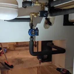 Fragment kotłowni w trakcie prac: widoczne elementy instalacji, rury w izolacji, manometr, pompa obiegowa LFP, narzędzia (wiertarka Milwaukee, klucz do rur) oraz akcesoria na podłodze z płytek...