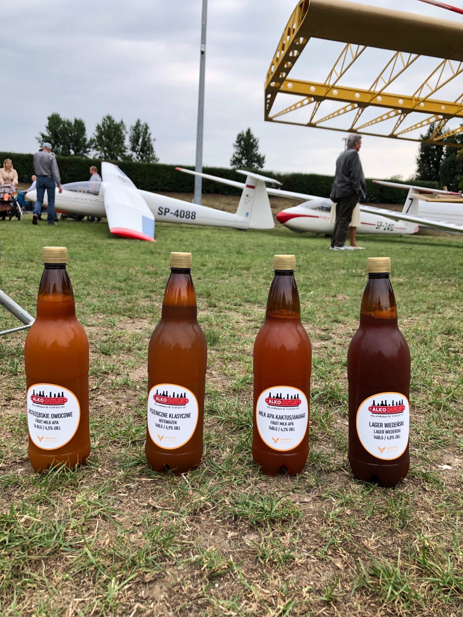Cztery butelki piwa rzemieślniczego różnych smaków, ustawione na trawie, z szybowcami w tle i osobami spacerującymi w oddali.