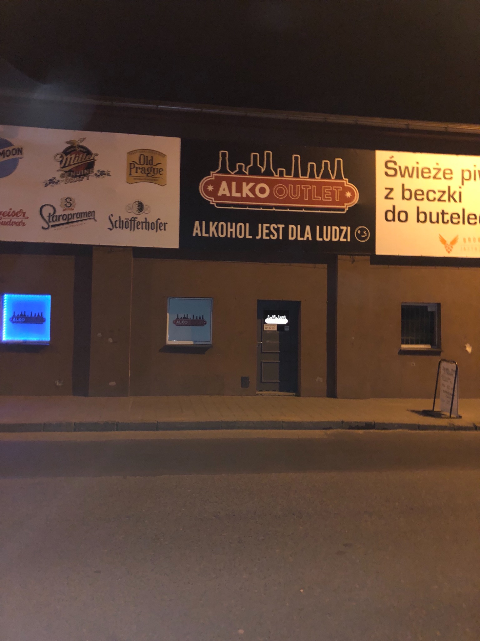 Fasada budynku z szyldem 'Alko Outlet' i reklamami różnych marek piwa, oświetlona w nocy, z chodnikiem na pierwszym planie.