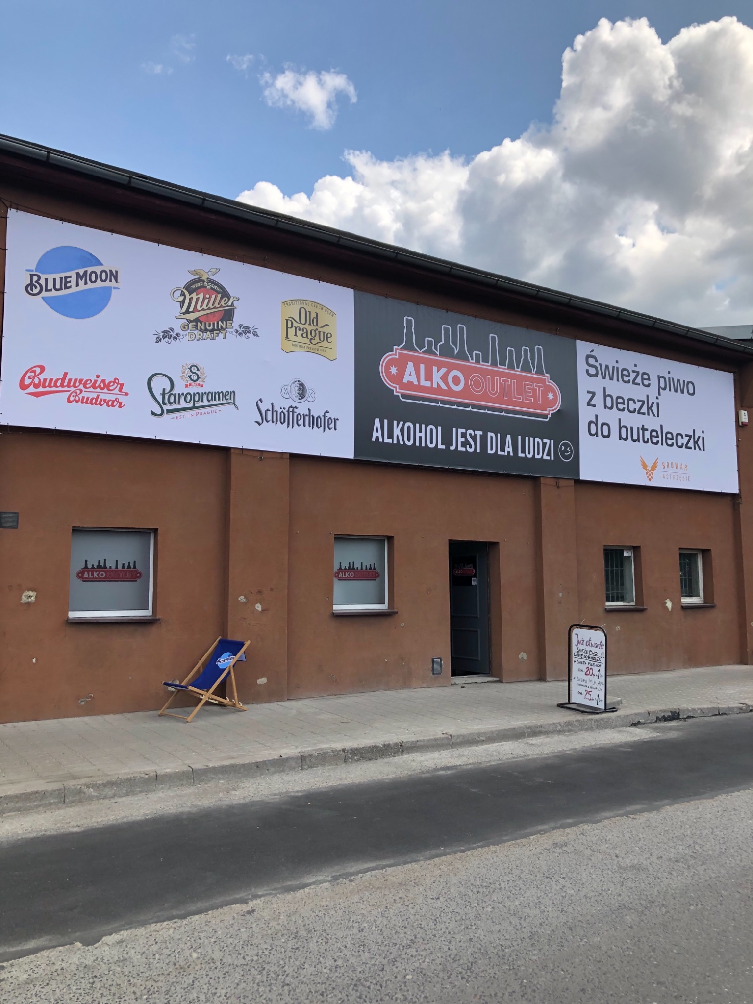 Fasada budynku z reklamami piw różnych marek i napisem 'ALKO OUTLET, ALKOHOL JEST DLA LUDZI', z leżakiem reklamowym Blue Moon i tablicą informującą o cenach, widok z ulicy.