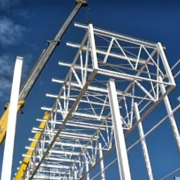 Montaż białej konstrukcji stalowej hali z użyciem żółtego dźwigu na tle błękitnego nieba.