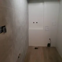 Łazienka w trakcie remontu: ściany wyłożone brązowymi płytkami, przygotowane otwory na gniazdka, sufit z płyt kartonowo-gipsowych, widoczny odpływ liniowy w podłodze.