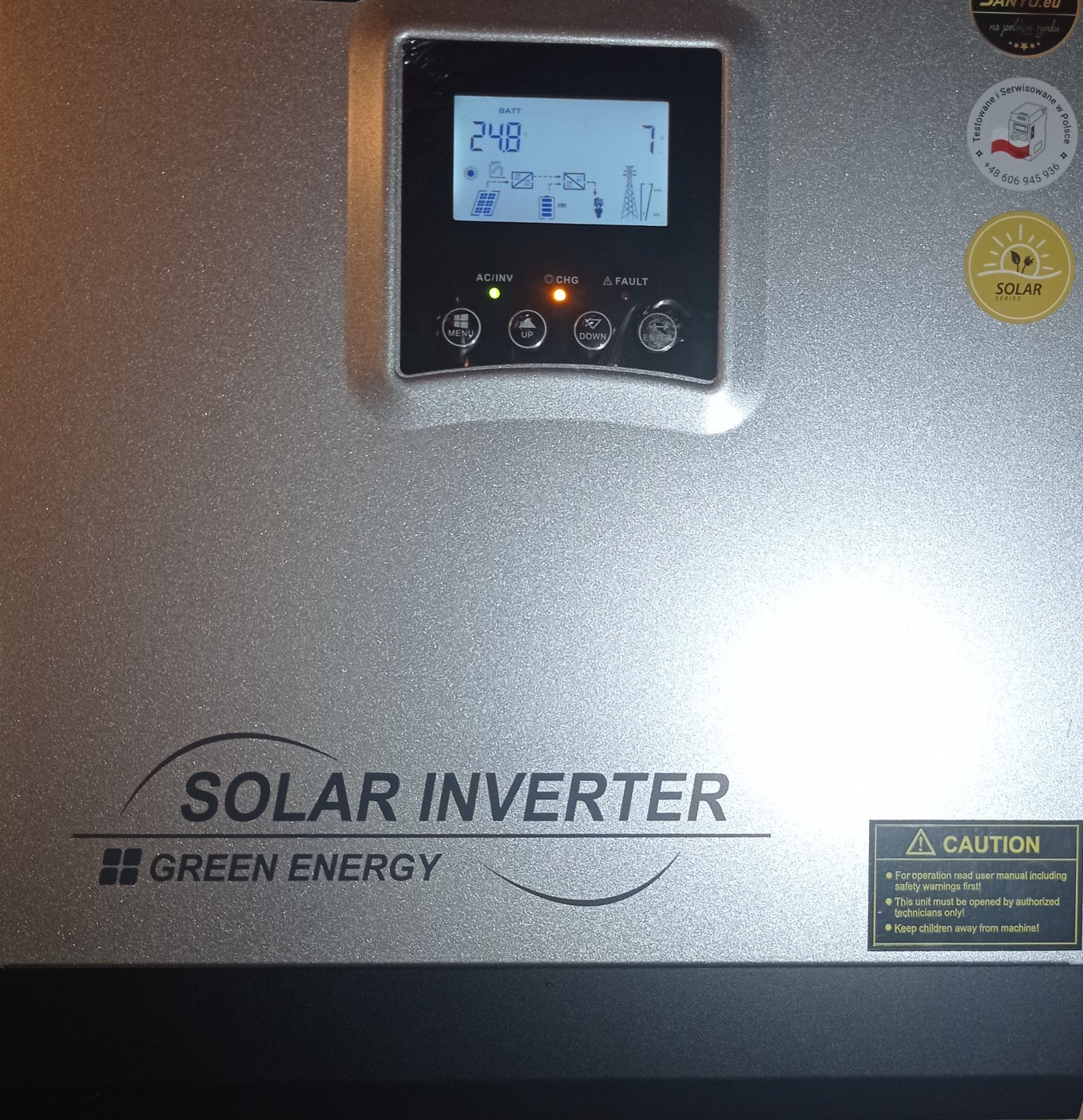 Szary inwerter solarny z wyświetlaczem pokazującym stan baterii i schemat przepływu energii, z naklejkami informacyjnymi i ostrzegawczymi na obudowie.