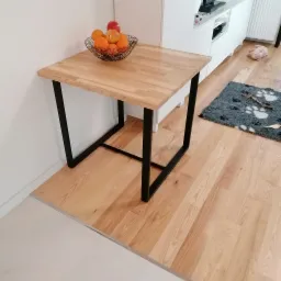 Drewniany stół z blatem z jasnego drewna i czarnymi, metalowymi nogami, na blacie druciany koszyk z owocami, w tle fragment kuchni.