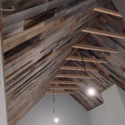 Skośny sufit wykończony panelami z postarzanego drewna, widoczne elementy konstrukcyjne i wiszące przewody oświetleniowe, wnętrze w trakcie prac wykończeniowych.