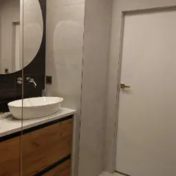 Nowoczesna łazienka z okrągłym lustrem, białą umywalką nablatową, szafką z drewnopodobnym fornirem, szarą płytką imitującą kamień na ścianach i białą, minimalistyczną stolarką drzwiową.