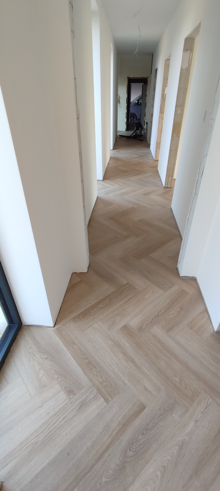 Korytarz z ułożonymi panelami w jodełkę, białe ściany w trakcie wykańczania, widoczne surowe wnęki drzwiowe i fragment okna po lewej.