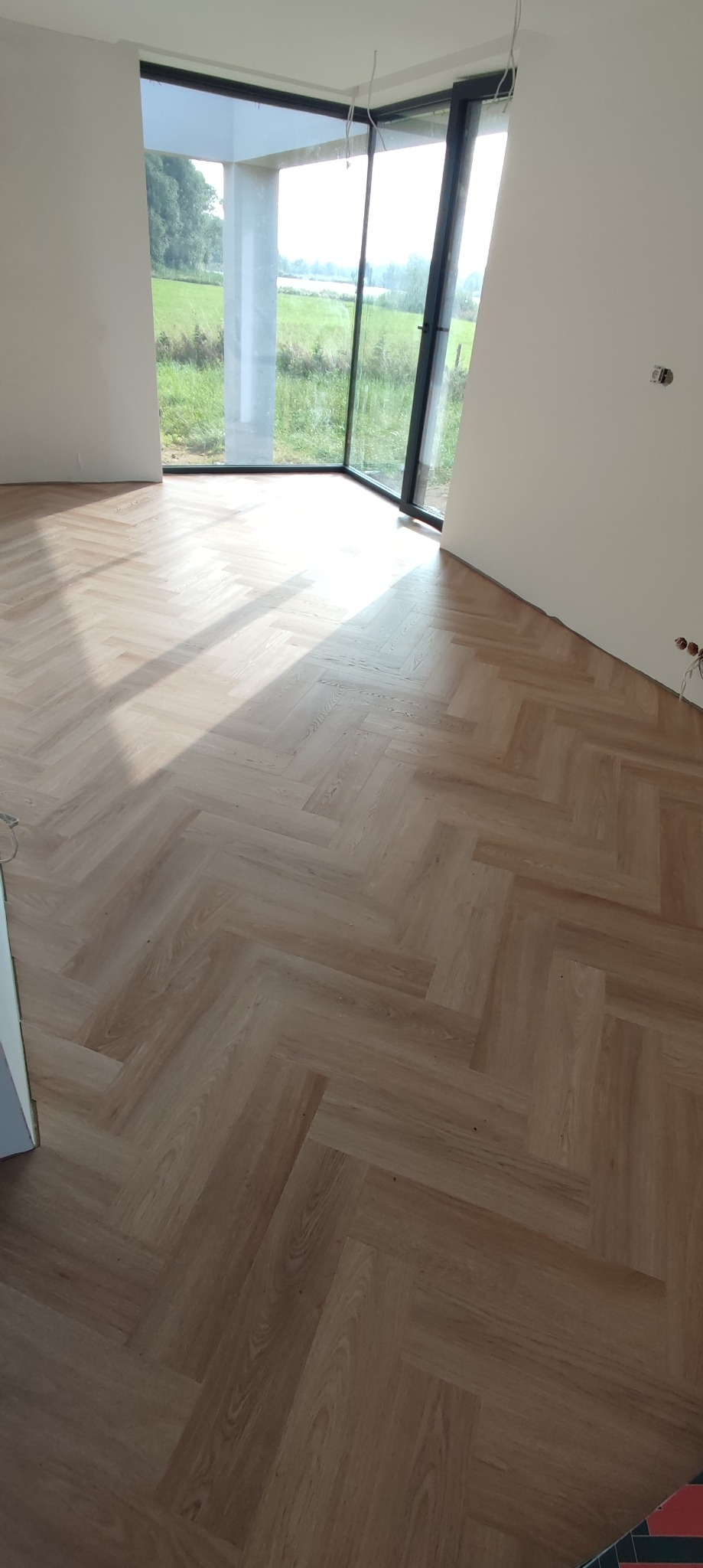 Jasne panele ułożone we wzór jodełki francuskiej w pustym pokoju z dużym oknem narożnym z widokiem na zielony krajobraz.