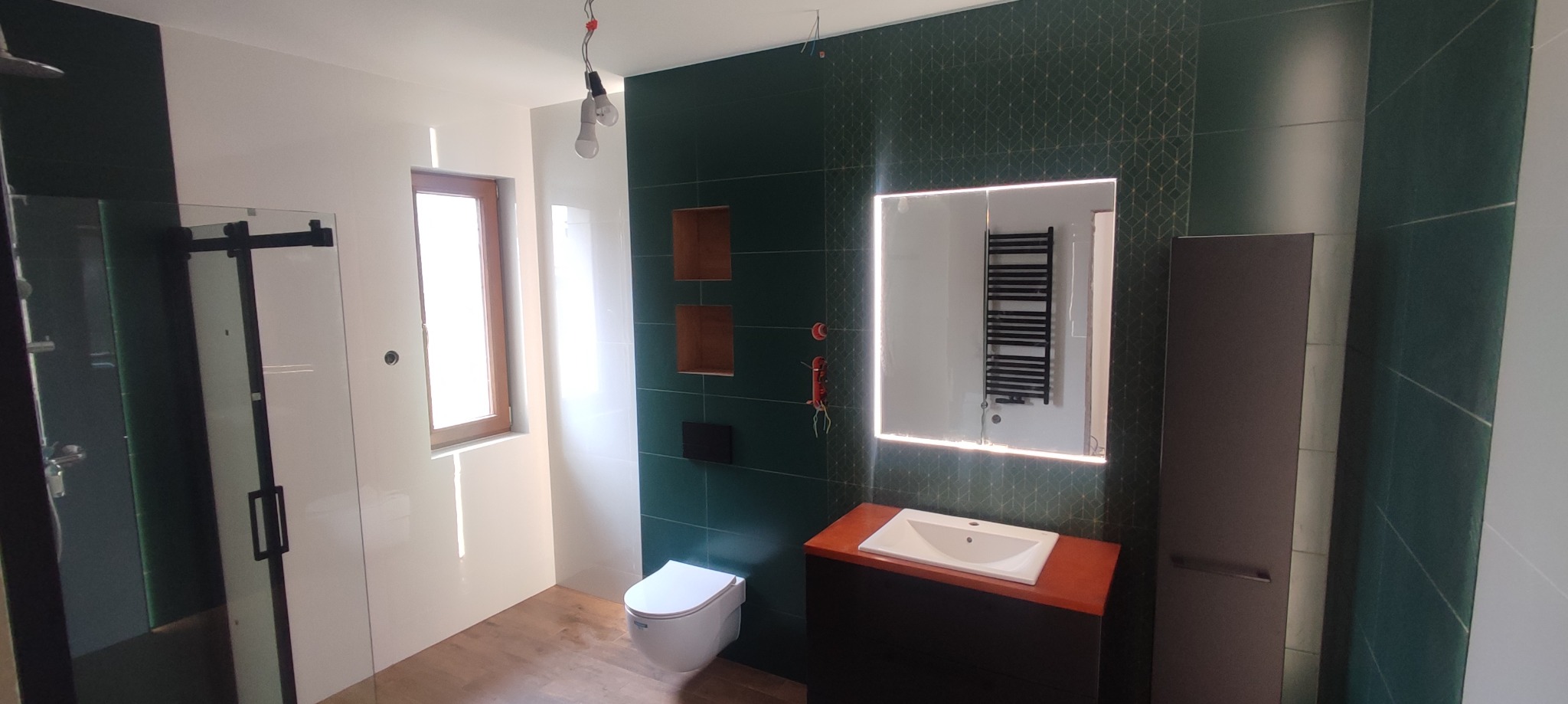 Nowoczesna łazienka w trakcie wykańczania: zielone płytki z geometrycznym wzorem, drewniana podłoga, biała toaleta i umywalka z szafką, kabina prysznicowa z czarnymi drzwiami przesuwnymi...