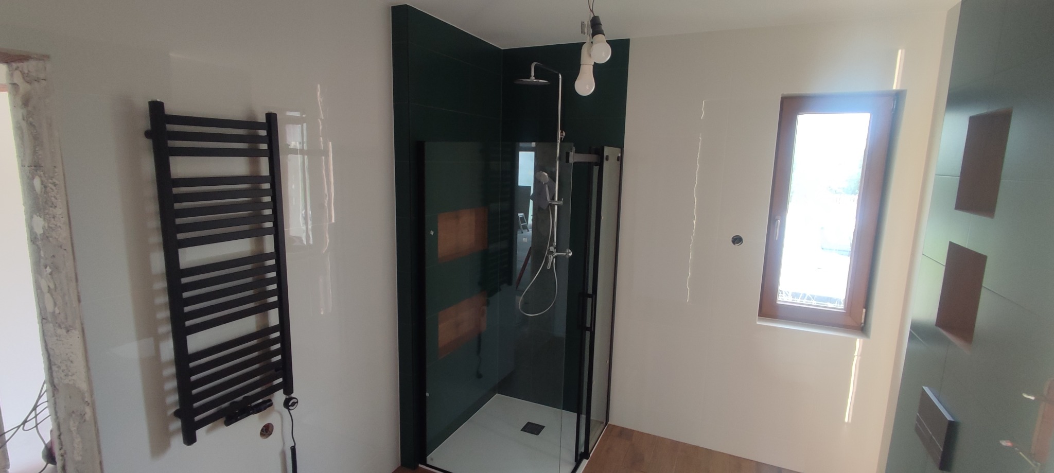 Nowoczesna łazienka z kabiną prysznicową typu walk-in z czarnymi profilami, grzejnikiem drabinkowym i fragmentem ściany z ciemnozielonych płytek z drewnianymi akcentami. Widoczne surowe wykończenia...
