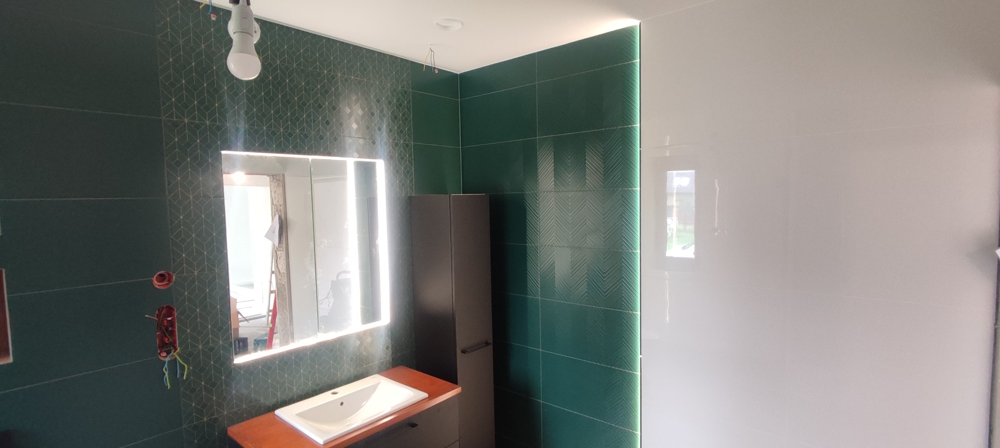 Nowoczesna łazienka w trakcie wykańczania: zielone płytki z geometrycznym wzorem, białe gładkie płytki, podświetlane lustro nad umywalką z szafką, widoczne niedokończone instalacje elektryczne.
