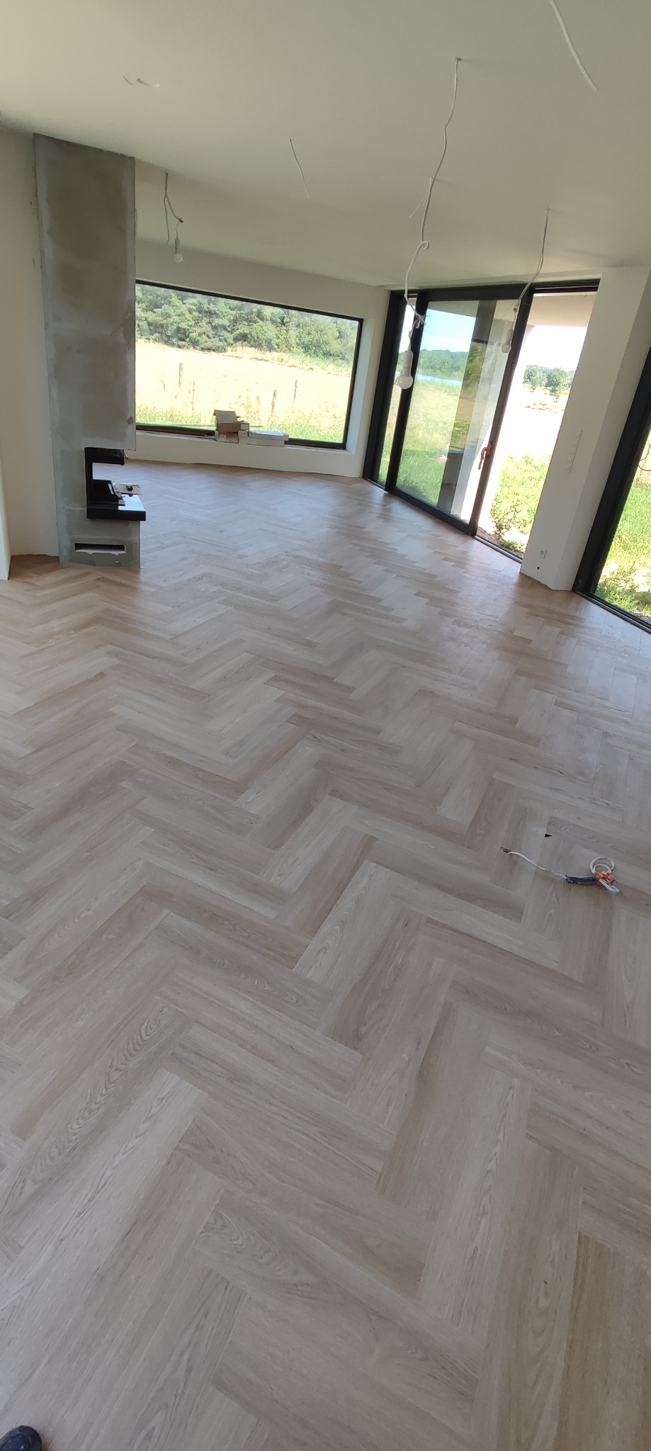 Jasna podłoga ułożona w jodełkę w przestronnym, jasnym pokoju z dużymi oknami i betonowym kominkiem w trakcie wykończenia, narzędzia do cięcia paneli leżą na podłodze.