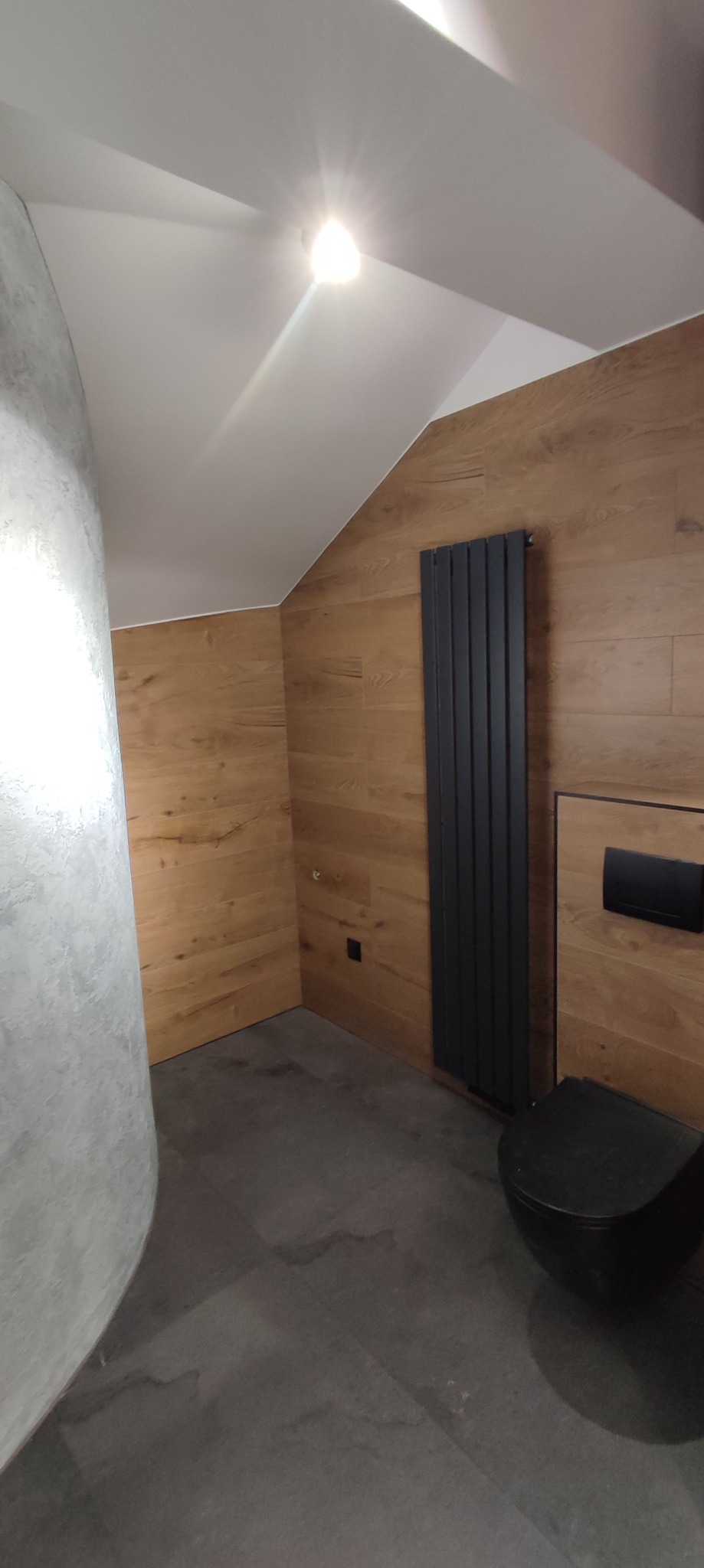 Nowoczesna łazienka w odcieniach drewna i szarości z czarną armaturą, grzejnikiem panelowym i podwieszaną toaletą w pomieszczeniu ze skośnym sufitem.