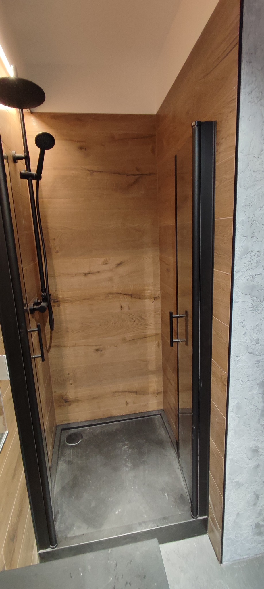 Kabina prysznicowa z czarną armaturą i drewnopodobnymi płytkami na ścianach, widoczny brodzik z odpływem liniowym.
