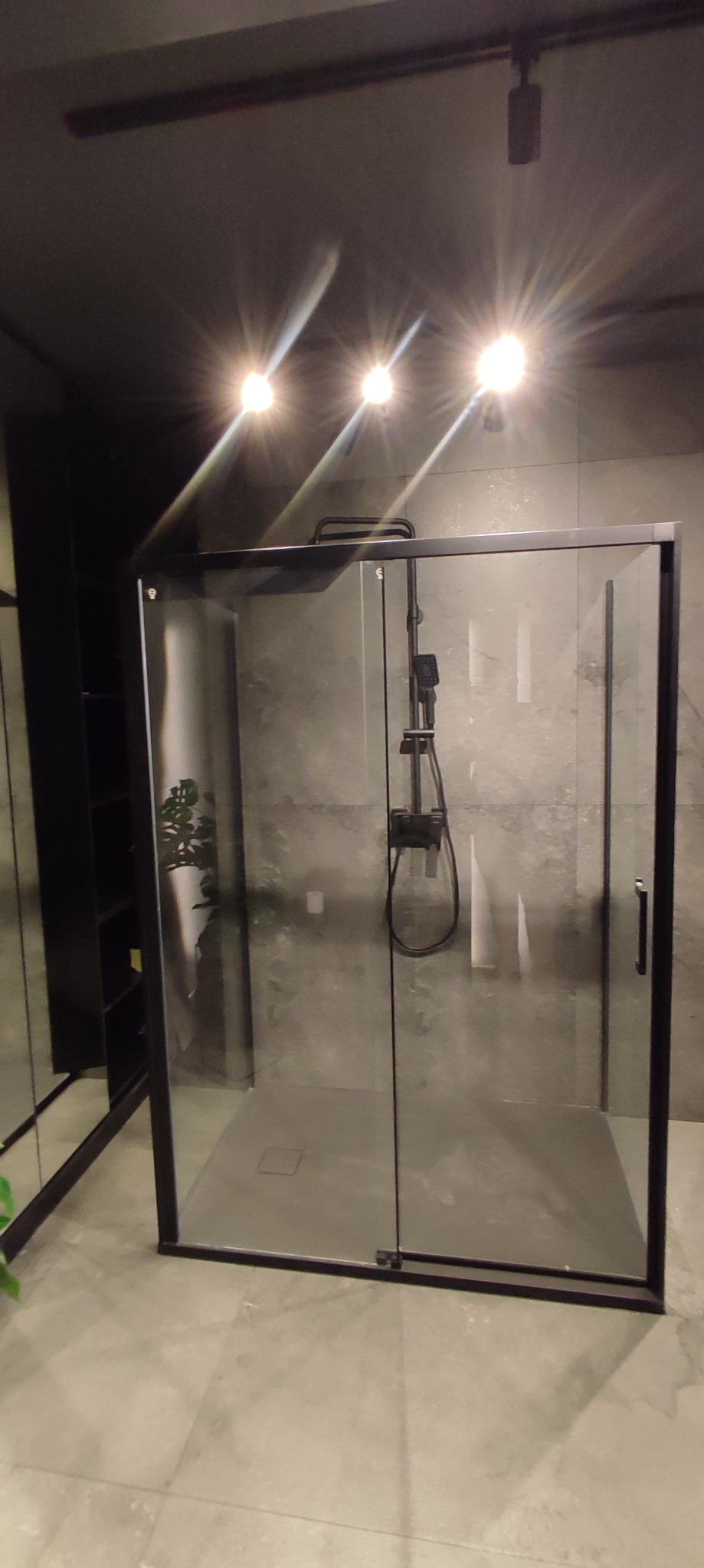 Nowoczesna kabina prysznicowa z czarnymi profilami i deszczownicą, szare płytki na ścianach, oświetlenie punktowe w suficie, widoczny fragment szafy z półkami.