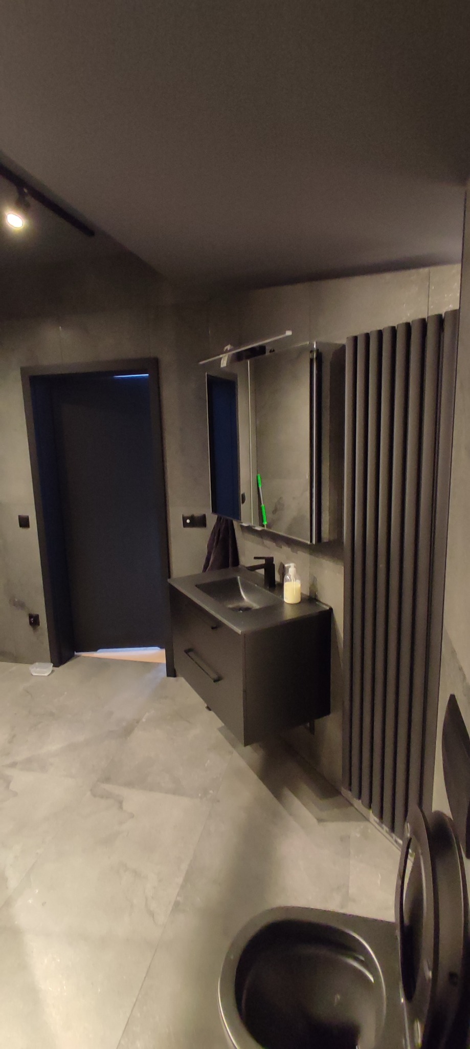 Nowoczesna łazienka w odcieniach szarości, z czarną umywalką nablatową, wiszącą szafką, grzejnikiem żeberkowym i lustrem z oświetleniem, wykończona płytkami imitującymi kamień.