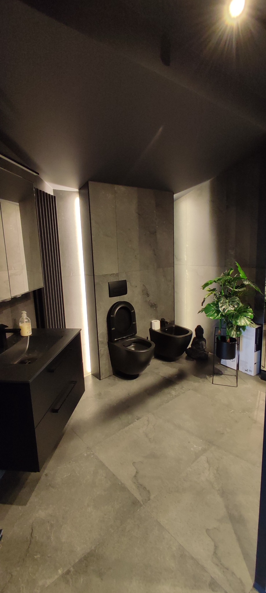 Nowoczesna łazienka z czarną armaturą: toaleta, bidet i umywalka na tle szarych płytek i dekoracyjnej zieleni w doniczce.