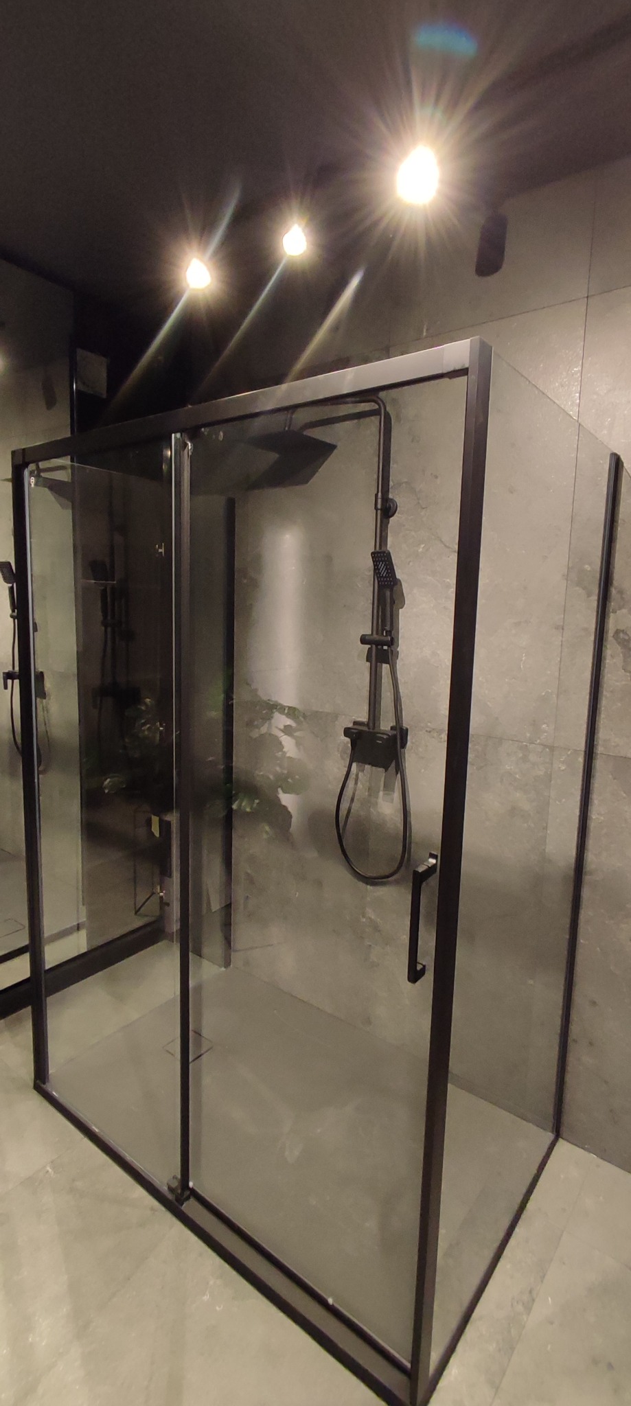Nowoczesna kabina prysznicowa z czarnymi profilami, deszczownicą i słuchawką, szara płytka imitująca kamień na ścianach i podłodze, oświetlenie punktowe sufitowe.