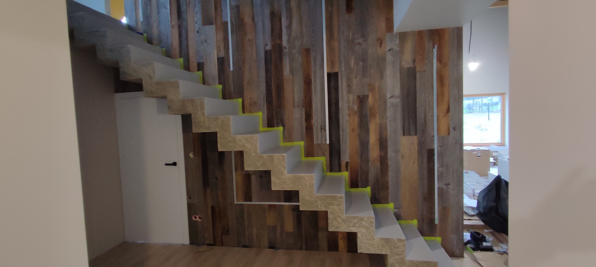 Nowoczesne schody dywanowe z okładziną imitującą kamień, zabezpieczone żółtą taśmą malarską, na tle ściany wykończonej drewnem z odzysku.