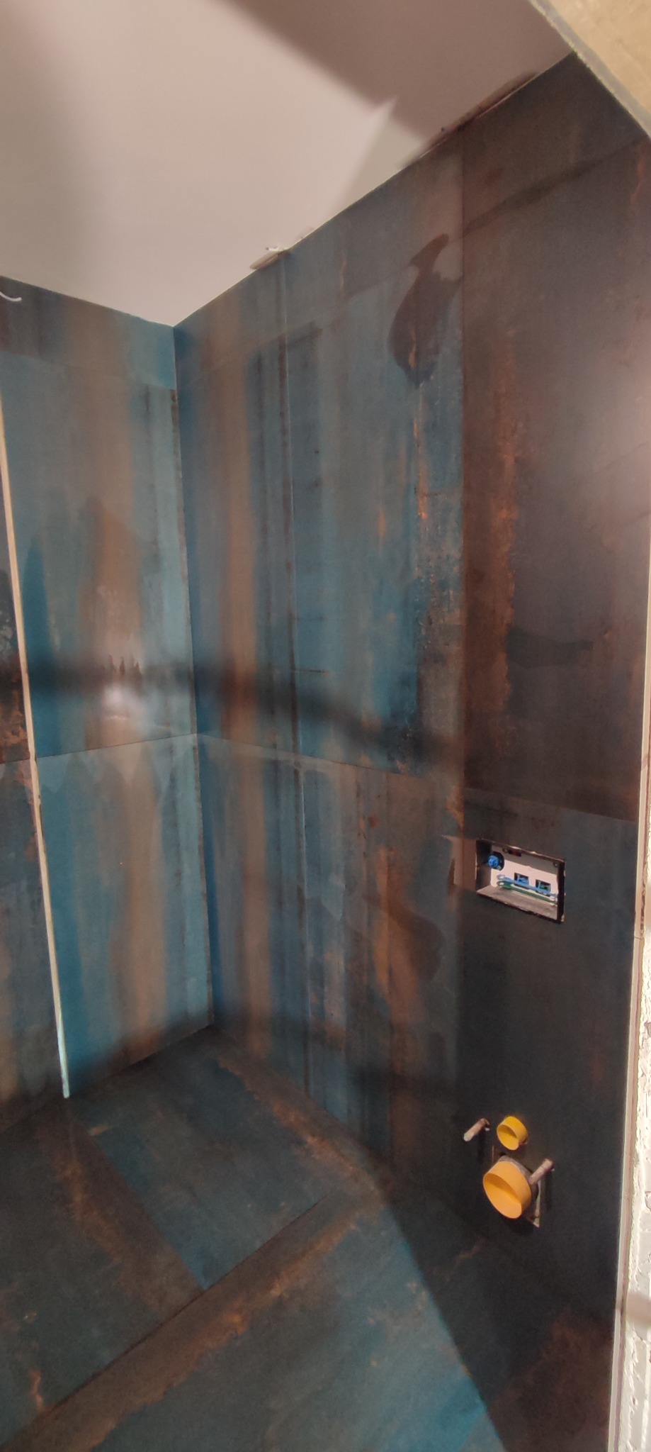 Narożnik kabiny prysznicowej z wykończonymi ścianami i podłogą w płytkach imitujących rdzę, widoczne wyprowadzenia instalacji wodnej i elektrycznej.