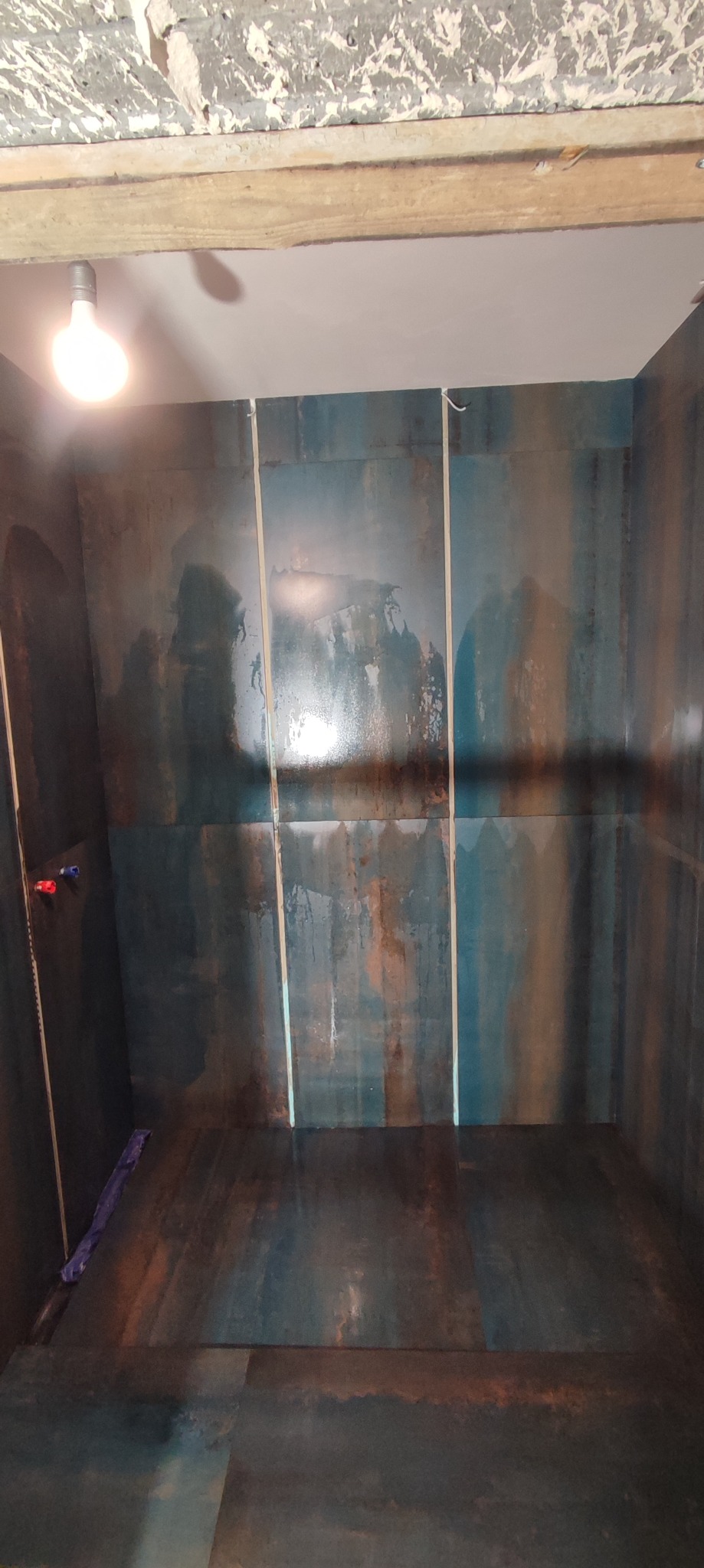 Wnętrze kabiny prysznicowej z ciemnymi, wzorzystymi płytkami na ścianach i podłodze, widoczne elementy instalacji wodnej, surowy strop z drewnianą belką i resztkami tynku.