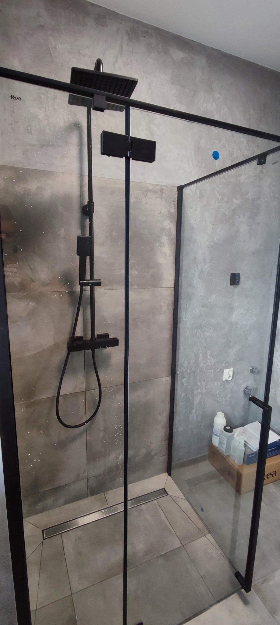 Nowoczesna kabina prysznicowa z czarnymi profilami, deszczownicą i odpływem liniowym, szare płytki imitujące beton i butelki środków czystości w rogu.