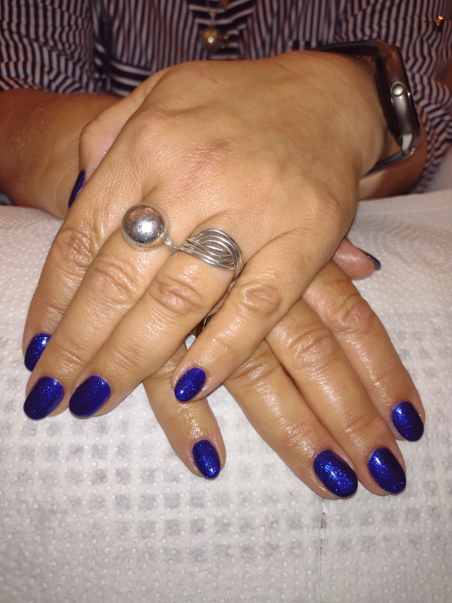 Dłonie z ciemnoniebieskim, błyszczącym manicure, srebrny pierścionek z kulką i zegarek na nadgarstku, na białym tle.